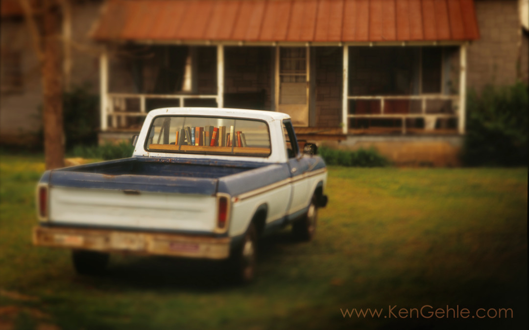 Old truck parked in front of a cabin with a rusted tin roof. There are books on a shelf in the window where a gun rack would be.